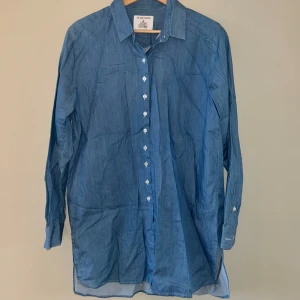 Skjorta från The Shirt Factory  - Tunn, blå skjorta från The Shirt Factory. Lite längre modell. Små slitsar på sidorna. 