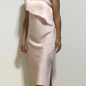 Ljusrosa klänning med fransdetalj från MOTF - Säljer en superfin ljusrosa klänning från MOTF. Den har en unik fransdetalj över bröstet som ger en cool touch. Klänningen är ärmlös och har en slits baktill för extra rörelsefrihet. Perfekt för festliga tillfällen!