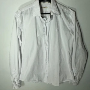 Vit skjorta från Burberry Brit - Säljer en stilren vit skjorta från Burberry Brit med klassiskt rutigt mönster på insidan av kragen och ärmsluten. Skjortan har långa ärmar och knappar framtill. Storlek L men skulle säga att den sitter som M 