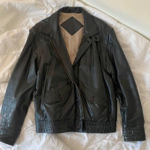 Svart skinnjacka med dragkedja - Snygg svart skinnjacka med dragkedja och knappar. Jackan har långa ärmar och flera praktiska fickor. Insidan är fodrad med ett randigt tyg för extra komfort. Perfekt för en cool och stilren look.