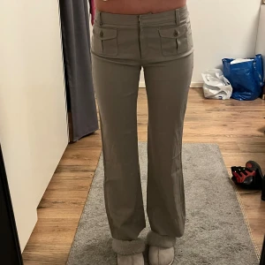 Beige byxor med fickdetaljer - Snygga beige byxor med låg/mid midja och bootcut-stil. Byxorna har framfickor med knappar och en stilren design som passar perfekt för en trendig look. Jag är 174 och dem passar exakt i längden. De är liknande kostymbyxor 