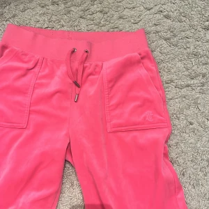 Rosa mjukisbyxor från Juicy Couture - Snygga rosa mjukisbyxor från Juicy Couture med snörning i midjan och praktiska fickor både fram och bak. Perfekta för en avslappnad stil med en touch av glamour.