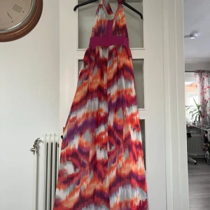 Klänning - Säljer en färgglad långklänning med ett abstrakt mönster i lila, orange,rött,grått och vitt. Klänningen har en halterneck-design med knytband och en bred lila midja som ger en smickrande passform. Perfekt för sommarens festligheter! Öppen rygg! Storlek S men passar även M perfekt!🥂🪻🌺💋Nytt skick. Kontakta mig för fler bilder eller prisförslag!