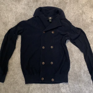 Mörkblå kofta från H&M - Säljer en stilren mörkblå kofta från H&M med dubbelknäppning och bred krage. Perfekt för kyligare dagar och ger en elegant touch till din outfit. Koftan har långa ärmar och ribbade muddar.