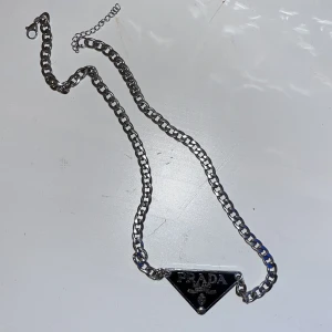 Silverfärgat halsband från Prada - Stilrent silverfärgat halsband från Prada med en kedja och en triangulär platta med logotyp. Perfekt för att ge en lyxig touch till din outfit. Skön under sommaren säljs billigt för har inget kvitto men är helt äkta.
