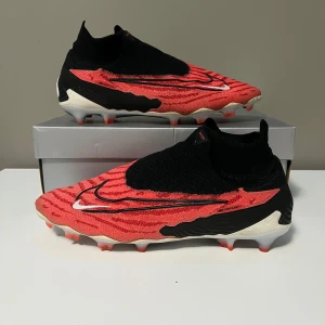 Nike Phantom GX fotbollsskor - Snygga Nike Phantom GX fotbollsskor i röd och svart med vit logga. Skorna har en strumpliknande passform och en unik mönstrad design. Perfekta för fotbollsplanen med sina greppvänliga dobbar. Om du vill ha fler bilder så är det bara att skicka!. (Priset kan diskuteras!)
