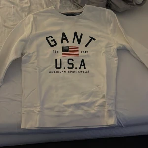 Vit sweatshirt från GANT - Säljer en vit sweatshirt från GANT med tryck av amerikansk flagga och texten 'GANT USA'. Tröjan har rund hals och långa ärmar, perfekt för en avslappnad stil.
