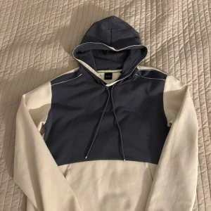 Grå och vit/beige hoodie från Boss - Snygg hoodie från Boss i grått och vitt/ beige med justerbar huva och dragsko. Perfekt för en avslappnad stil. Boss-loggan är diskret placerad på bröstet.