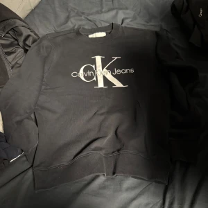 Svart sweatshirt från Calvin Klein Jeans - Säljer en stilren svart sweatshirt från Calvin Klein Jeans med deras ikoniska logga i vitt på framsidan. Tröjan har långa ärmar och ribbade muddar vid ärmslut och nederkant. Perfekt för en avslappnad look.