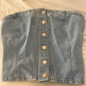 Jeans korsett med knappar - Snygg jeans korsett i ljusblå denim med knappar framtill. Perfekt för en trendig look. Baksidan har elastiska detaljer för bättre passform.