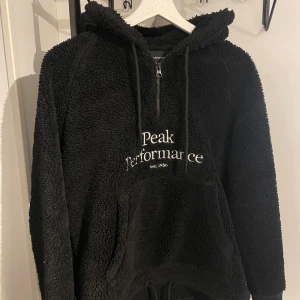 Svart Teddy (fleece)  hoodie från Peak Performance - Mysig svart Teddy hoodie från Peak Performance med dragkedja och känguruficka. Perfekt för kyliga dagar med sin varma och mjuka känsla. Stort tryck på framsidan med märkesnamnet.