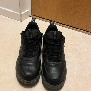 Svarta Nike Air Force sneakers - Säljer ett par svarta Nike Air Force sneakers med klassisk design och snörning. Skorna har en diskret Nike-logga i blått på sidan och är tillverkade i skinn.