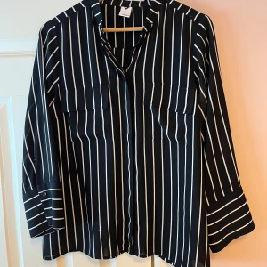 Svartvit randig blus - Snygg svart blus med vita ränder och långa ärmar. Blusen har en avslappnad passform och knappar framtill. Perfekt för en stilren look.