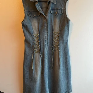 Jeansklänning från Boule - Snygg ärmlös jeansklänning från Boule med snörning framtill och två bröstfickor med knappar. Perfekt för en avslappnad stil med en touch av edge. Klänningen har en klassisk krage och knäppning hela vägen ner.