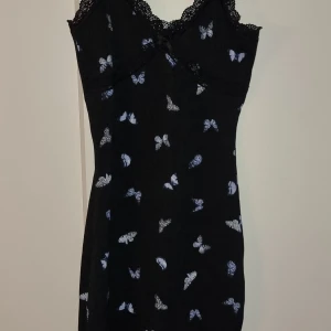 Svart klänning - Svart klänning från Divided med fjärilsmönster i blått och vitt. Klänningen har spetsdetaljer vid halsringningen och smala axelband. Sitter tight och snyggt på kroppen. Storleken är xxs men skulle passa xs och möjligtvis en liten s.