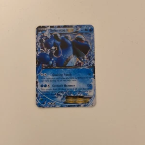Seismitoad EX Pokémonkort - Säljer ett Seismitoad EX Pokémonkort med kraftfulla attacker som Quaking Punch och Grenade Hammer. Kortet har en blå och silver design med en bild av Seismitoad. Perfekt för samlare eller spelare som vill förstärka sin kortlek.