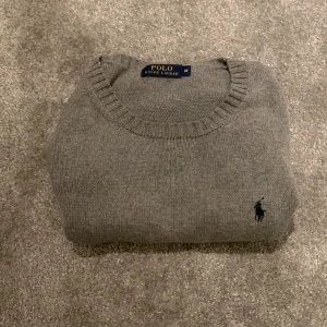 Grå tröja från Ralph Lauren - Säljer en stilren grå tröja från Ralph Lauren med rund hals och ribbade muddar. Den har en diskret broderad logga på bröstet. Perfekt för en klassisk och avslappnad look. Storlek M, skisk 8/10. Ny pris 1200kr