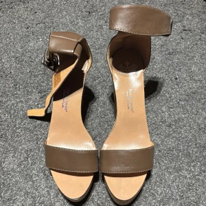 Bruna sandaler med kilklack - Snygga bruna sandaler med kilklack och justerbart spänne runt ankeln. Perfekta för sommarens alla tillfällen. Tillverkade i skinn med en bekväm passform.