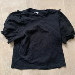 Svart kortärmad topp med puffärmar cubus - Snygg svart topp med korta puffärmar. Perfekt för en stilren look. Tillverkad i ett mjukt material som ger en bekväm passform.