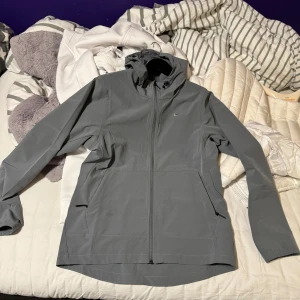 Grå vindjacka från Nike - Säljer en stilren grå vindjacka från Nike med huva och dragkedja. Jackan har en minimalistisk design med diskret logga på bröstet och praktiska fickor. Perfekt för blåsiga dagar.