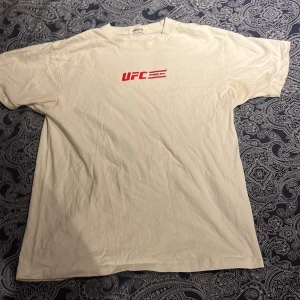 Vit UFC Dragon Ball t-shirt - Cool beige t-shirt med UFC och Dragon Ball-tryck. Framsidan har en röd UFC-logga, medan baksidan visar en stor Dragon Ball-illustration med texten 'GYM'. Lite oversized men go till gymmet eller casual.