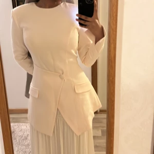 Beige/offwhite kavaj - Elegant beige/offwhite kavaj från med långärmad design och en stilren, figursydd passform. Klänningen har en unik asymmetrisk knäppning framtill och unika ärmar. Perfekt för en sofistikerad look, till event, jobbet eller till och med studenten. Skulle säga att den är stor i storlek så den rekommenderas för s/m. Aldrig använd!!! 