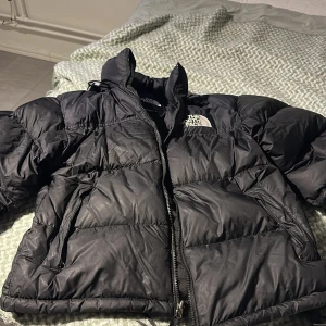 Svart dunjacka från The North Face - Säljer en svart dunjacka från The North Face. Jackan har en pufferdesign med dragkedja och hög krage för extra värme. Perfekt för kalla dagar.