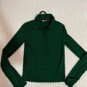 Grön ribbad tröja från Envii - Snygg grön ribbad tröja från Envii med knappar framtill. Tröjan har en klassisk krage och långa ärmar, perfekt för kyligare dagar. Den har en stilren design som passar till många olika outfits.