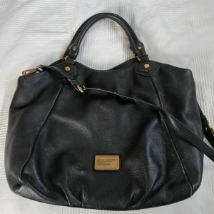 Svart väska  från Marc Jacobs - Snygg svart väska i skinn från Marc by Marc Jacobs med guldfärgade detaljer. Väskan har ett rymligt inre med ett mönstrat foder och en innerficka. Väskan är i väldigt fint skick men har lagt in bilder där det är slitet men det är absolut ingenting som syns. Priset kan diskuteras