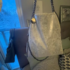 Silver axelväska med kedjedetaljer - Snygg silverfärgad axelväska med kedjedetaljer och glittrig yta. Väskan har en stilren design med en kedja som axelrem och en praktisk storlek för vardagsbruk. Perfekt för att ge din outfit en extra touch av glamour. Köpte väskan i grann canaria original pris 600kr