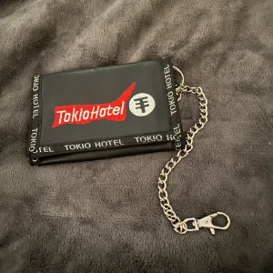 Svart plånbok med kedja från Tokio Hotel - Cool svart plånbok med kedja och Tokio Hotel-logga. Perfekt för fans av bandet. Plånboken har en stilren design med en praktisk kedja för extra säkerhet.
