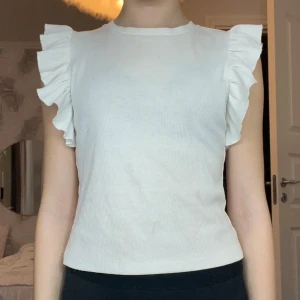 Vit topp med volangkortärmar - Säljer en stilren vit ribbad topp med volangdetaljer vid ärmarna. Perfekt för en snygg och enkel look. Toppen är ärmlös och har en snygg passform. Den kommer från SHEIN. Den kostade 100 kr när jag köpte den. Den är använd runt 5 gånger men är i väldigt bra skick. Den är jätte snygg och skön! 😍
