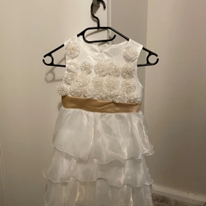 Vit ärmlös klänning med volanger - Charmig vit klänning med ärmlös design och vackra volanger. Överdelen är dekorerad med blommiga detaljer och ett elegant guldigt band i midjan. Perfekt för speciella tillfällen.