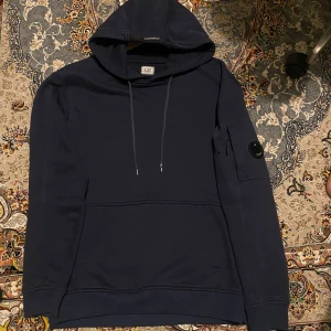 Mörkblå hoodie från C.P. Company - Säljer en stilren mörkblå hoodie från C.P. Company. Tröjan har en klassisk design med huva och dragsko samt en praktisk ficka på ärmen med knappdetalj. Jätte fin och bra shick pris kan diskuteras