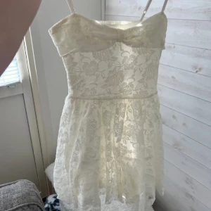 Vit spetsklänning med axelband - Charmig vit klänning i spets med smala axelband och en söt rosett framtill. Perfekt för sommarens festligheter med sin romantiska stil och korta längd.