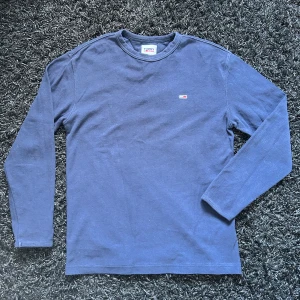 Blå långärmad tröja från Tommy Jeans - Säljer en stilren blå långärmad tröja från Tommy Jeans. Tröjan har en liten broderad logotyp på bröstet och är perfekt för en avslappnad look.