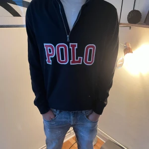 Svart tröja med dragkedja från Polo Ralph Lauren - Snygg svart tröja från Polo Ralph lauren  med stor logga i rött och vitt på framsidan. Tröjan har en dragkedja vid halsen och långa ärmar. Perfekt för en avslappnad stil.
