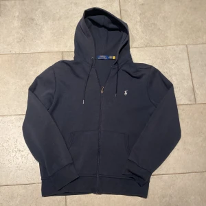 Ralph Lauren zip hoodie blå - Säljer en stilren mörkblå hoodie från Ralph Lauren med dragkedja och klassisk logga på bröstet. Perfekt för en avslappnad look. Den har en praktisk huva och fickor framtill.