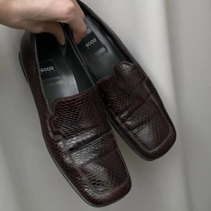 Bruna loafers från Ecco - Snygga bruna loafers från Ecco med ett elegant ormskinnsmönster. Skorna har en klassisk design med fyrkantig tå och är perfekta för en stilren look.