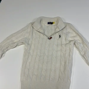 Beige stickad tröja från Ralph Lauren - Säljer en stilren vit stickad tröja från Ralph Lauren med kabelstickat mönster. Tröjan har en dragkedja vid halsen och långa ärmar samt en fungerande qr kod. Storleken är M men passar mera som S, skriv till mig vid frågor samt så kan pris diskuteras 