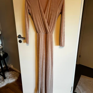 Beige omlottklänning från NLY Trend - Elegant beige långklänning från NLY Trend med omlottdesign och långa ärmar. Perfekt för en stilren look med sin mjuka och glittriga finish. Oanvänd men fattas prislappen 