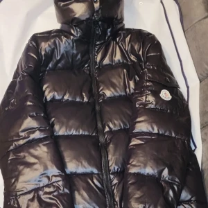 Svart dunjacka från Moncler - Säljer en snygg svart dunjacka från Moncler med hög krage och dragkedja. Jackan har en glansig finish och Moncler-logga på ärmen. Perfekt för kyliga dagar.