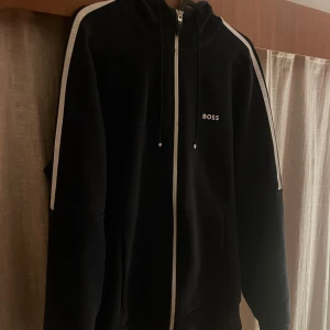 Svart hoodie från Hugo Boss - Snygg svart hoodie från Hugo Boss med dragkedja och vita detaljer längs ärmarna. Perfekt för en stilren och avslappnad look. Hoodien har en justerbar huva med snören och en diskret logga på bröstet. Oanvänd, inga defekter, priset går att diskutera.