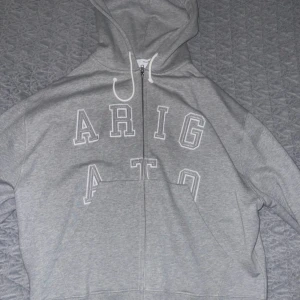 Grå hoodie från Axel Arigato - Säljer en stilren grå hoodie från Axel Arigato med dragkedja och texten 'ARIGATO' i vitt på framsidan. Tillverkad i ekologisk bomull, perfekt för en avslappnad look. Huvan har vita snören och insidan är mjuk och bekväm. Köpte för 3000 men säljer nu endast för 1200! Använd en gång så skicket är som nytt 