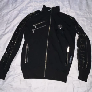 Philipp Plein x floyd maywheather - Snygg svart jacka från Philipp Plein med coola detaljer. Jackan har dragkedjor och ett märke på bröstet. På baksidan finns texten 'Floyd Mayweather' och 'Philipp Plein' i glittriga bokstäver. Perfekt för dig som vill sticka ut!