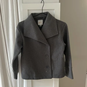 Grå kappa från Gina Tricot - Säljer en stilren grå kappa från Gina Tricot. Jackan har en klassisk design med stora knappar framtill och en bred krage. Perfekt för kyligare dagar och ger en elegant touch till din outfit.