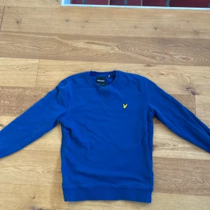 Blå tröja från Lyle & Scott - Säljer en snygg blå tröja från Lyle & Scott med deras ikoniska gula logga broderad på bröstet. Tröjan har långa ärmar och en rund halsringning. Perfekt för en stilren och avslappnad look.