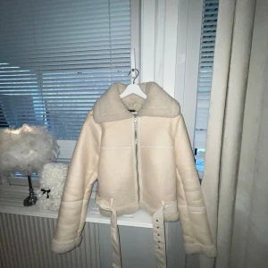 Beige teddyjacka med dragkedja - Säljer en mysig beige teddyjacka med dragkedja och bred krage. Jackan har långa ärmar och ett bälte i midjan för extra stil. Perfekt för kyliga dagar när du vill hålla dig varm och trendig. HELT OANVÄND