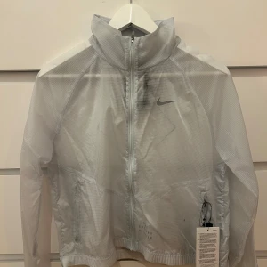 Vit vindjacka från Nike - Säljer en stilren vit vindjacka från Nike med dragkedja och hög krage. Genomskinlig, prefekt för träning
