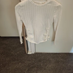 Vit stickad tröja - Säljer en vit stickad tröja med ett fint flätmönster. Tröjan har långa ärmar och en söt knytdetalj vid midjan. Perfekt för kyligare dagar!
